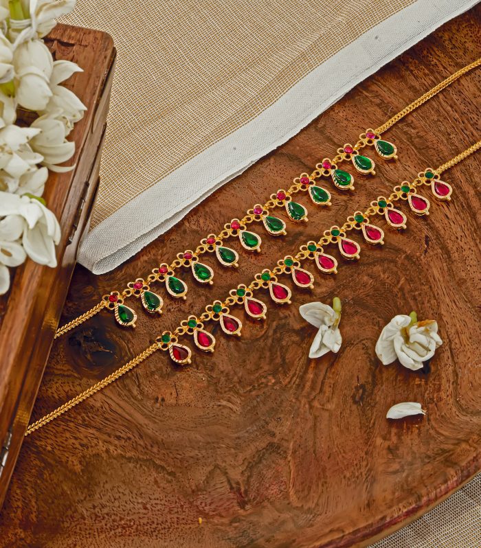 Two small traditional gold Nagapadam necklaces with alternating red and green teardrop stone pendants displayed on wooden surface.