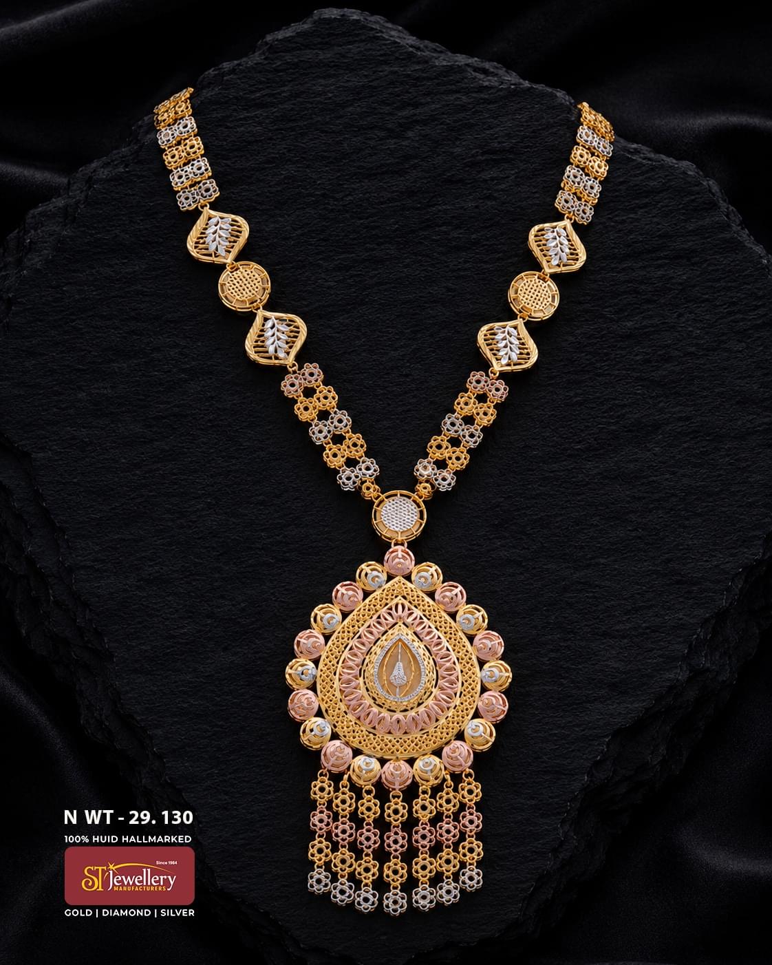 Multitone gold necklace with repeating heart and teardrop motifs, an ornate rose-gold floral pendant, white detailing, and cascading decorative drops displayed on a black background.