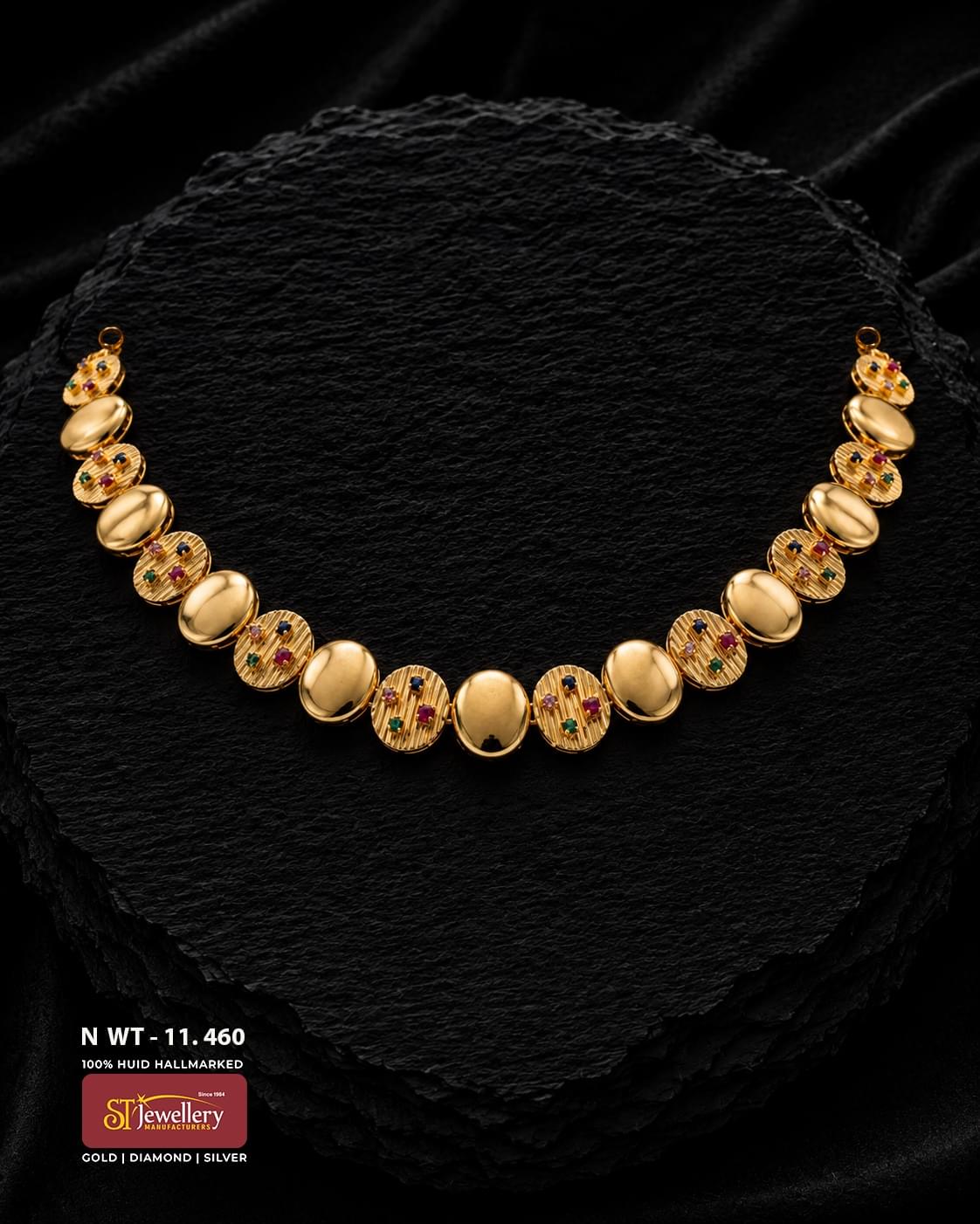 Gold necklace with alternating polished oval links and textured round motifs decorated with small red, green and dark-coloured stone accents, displayed on a black textured jewellery surface.
