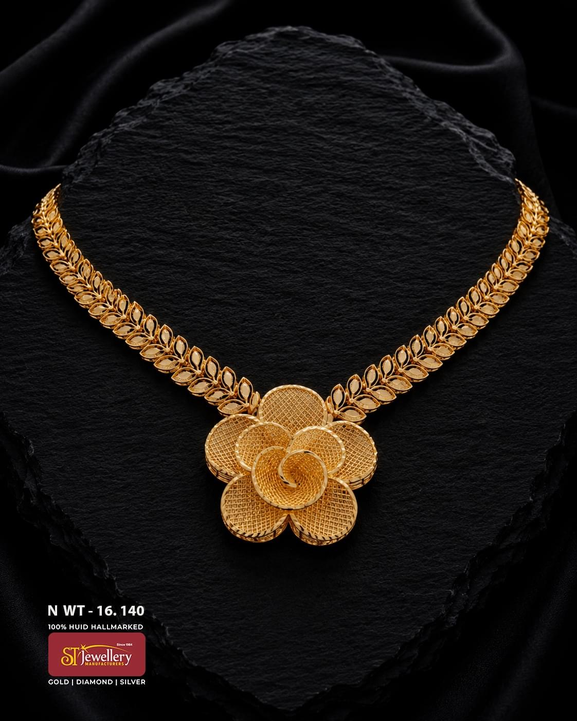 Gold necklace featuring a symmetrical leaf-pattern chain and large layered flower-shaped centrepiece with finely textured petals, displayed on a black textured background.
