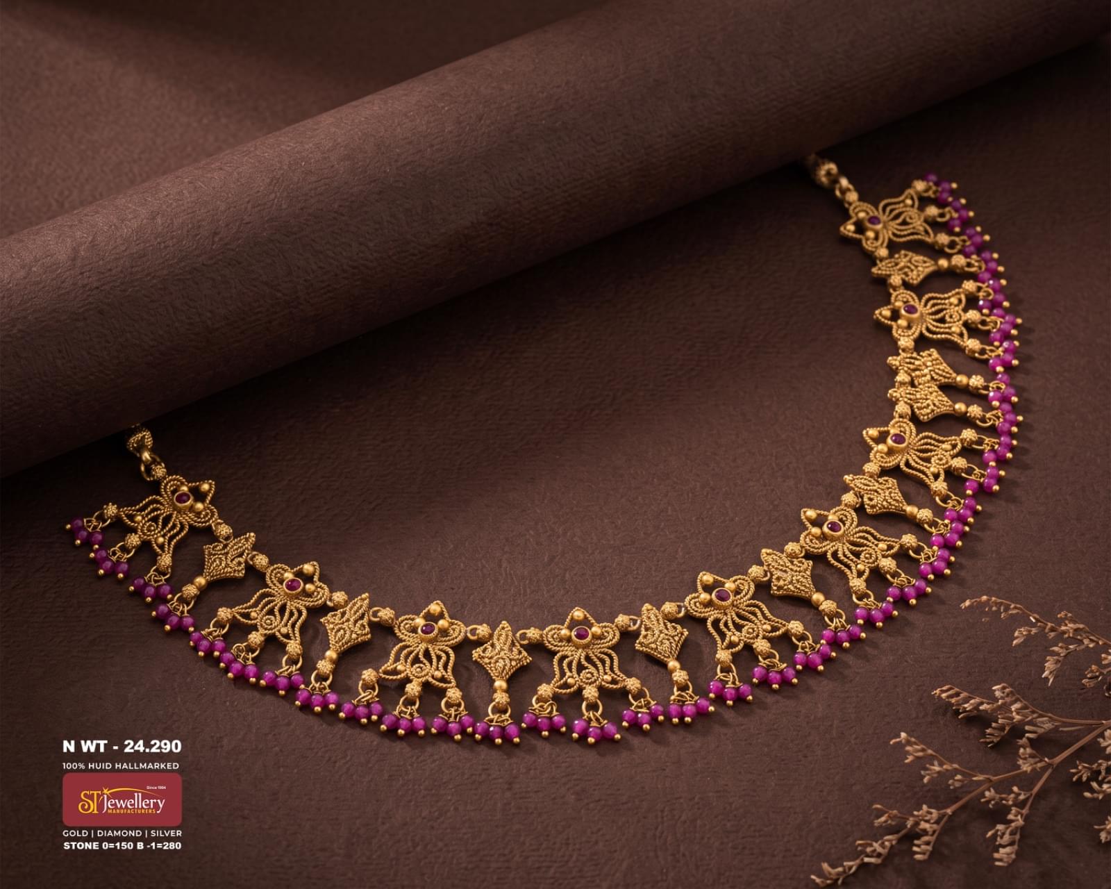 Antique gold necklace with intricate traditional motifs, ruby-coloured stone accents and a row of pink beads, displayed on a rich dark brown textured background.