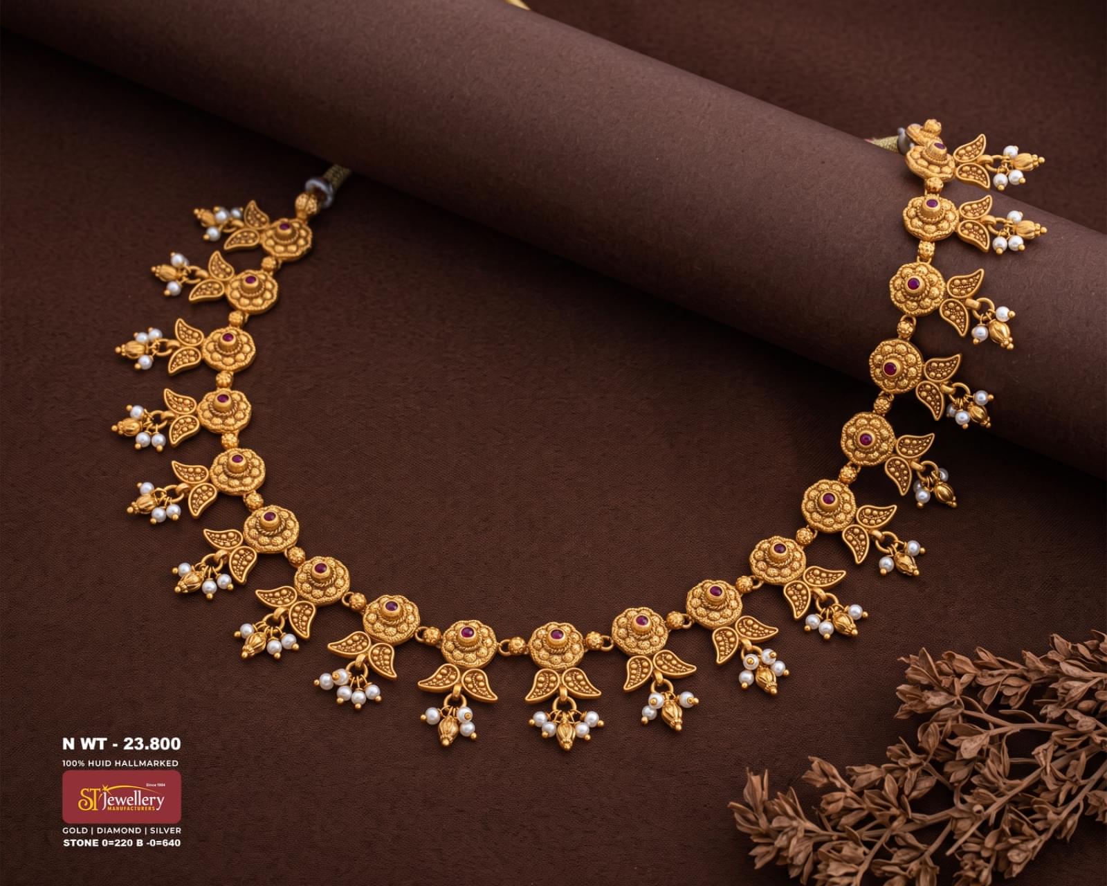 Antique gold necklace with floral motifs, red stone accents, leaf details and delicate white pearl drops, displayed on a rich brown textured background.